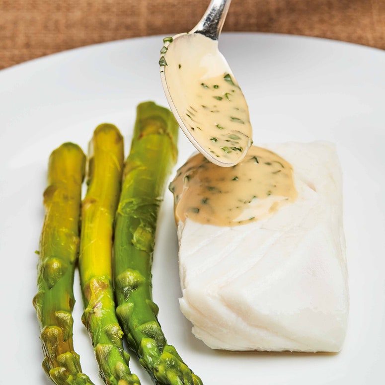 White plate with simple halibut fillet and spoon of herby vinaigrette sauce plus 3 asparagus spears alongside.